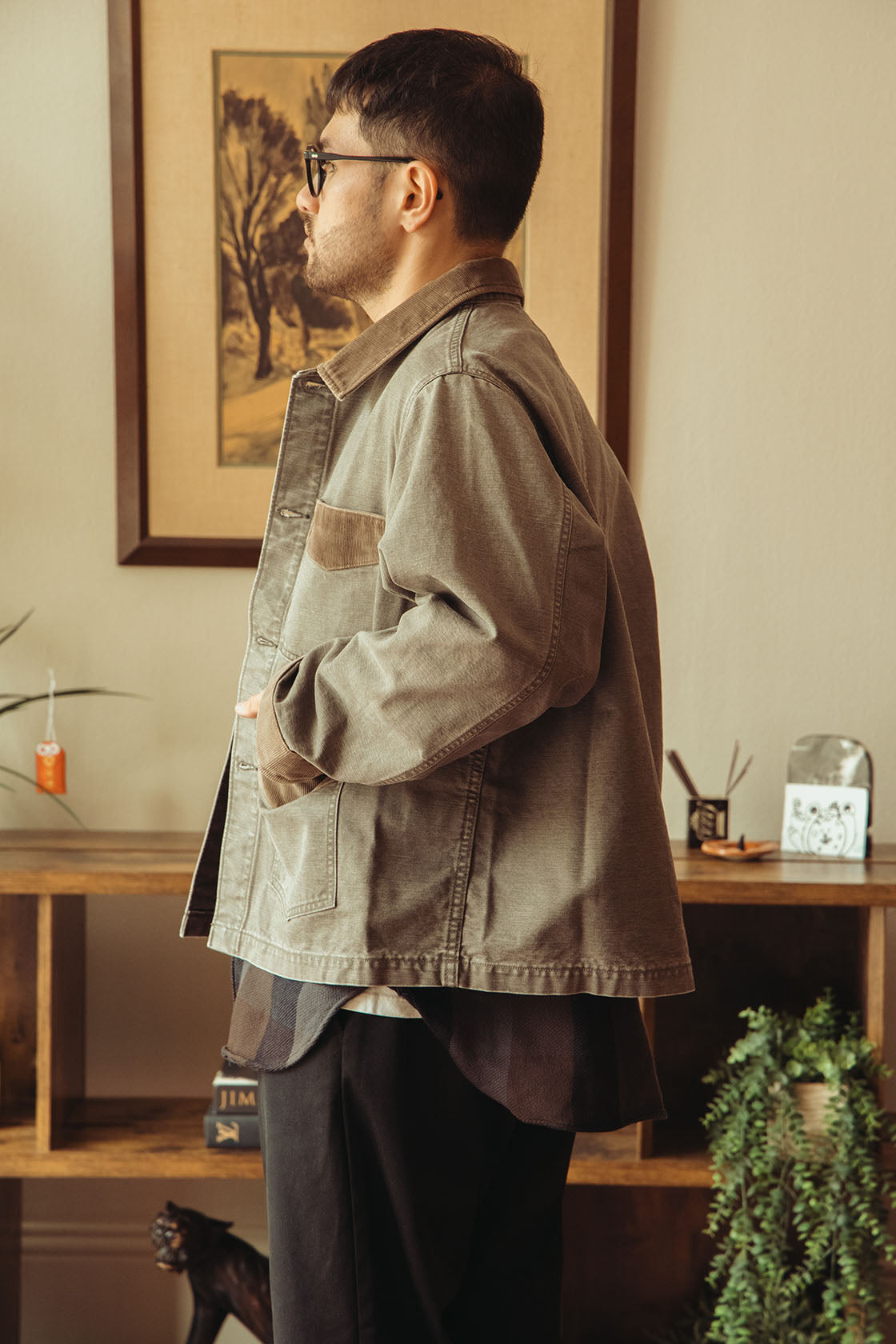 Profile shot of a man wearing a washed black jacket over a dark shirt and black pants, standing in a warm, indoor room with wooden shelves and framed art in the background.
