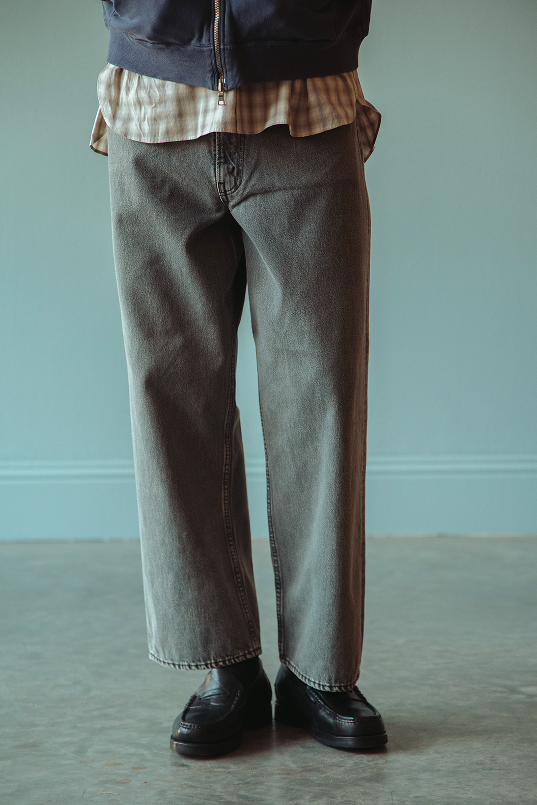 A person shown from the waist down wearing light gray wide-leg denim jeans, a navy blue sweatshirt layered over a tan plaid shirt, and black leather loafers, standing against a pale blue wall.
