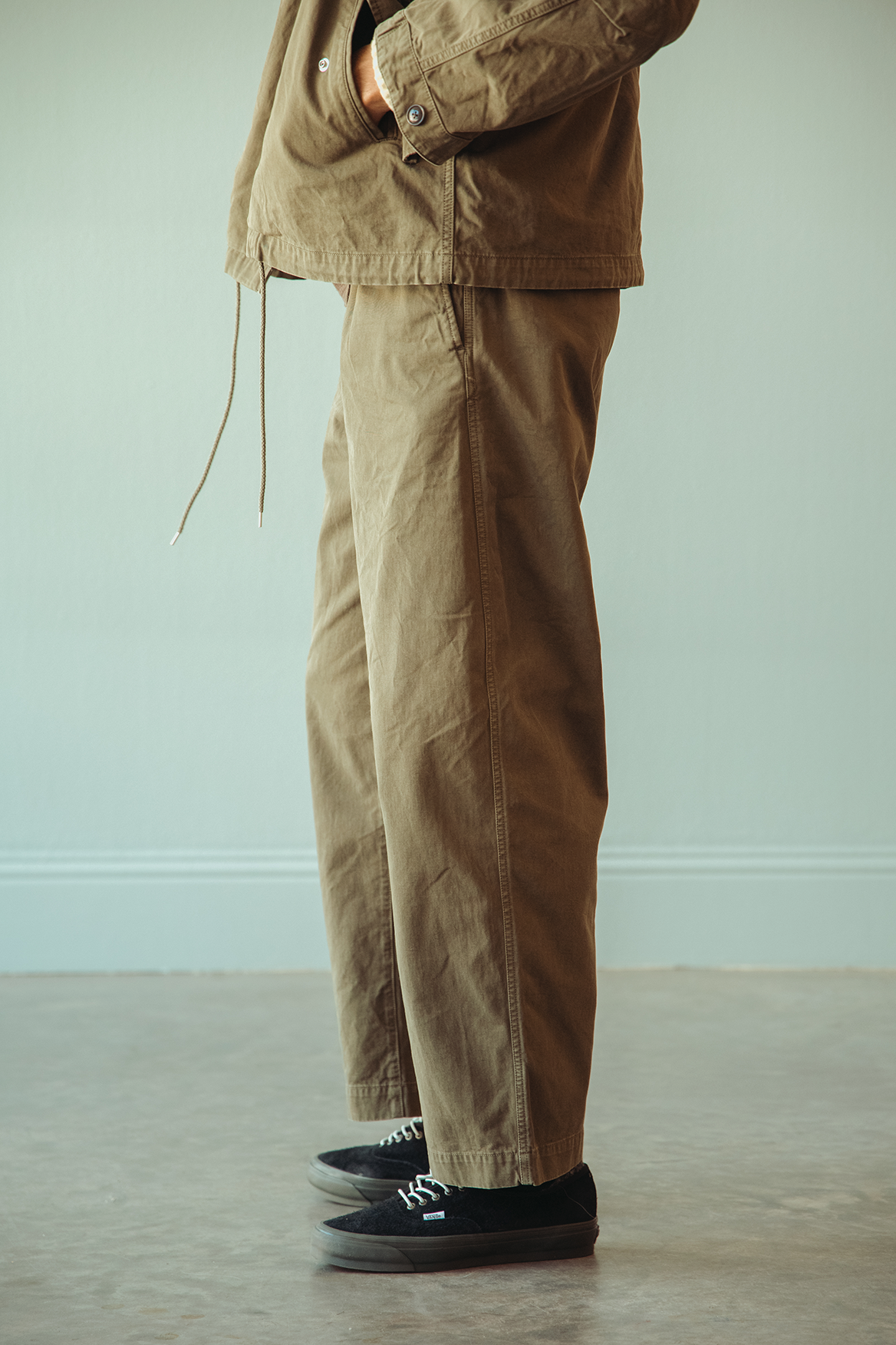 A person wearing loose brown utility pants and a matching brown jacket, standing indoors against a pale wall and concrete floor with black low-top sneakers.