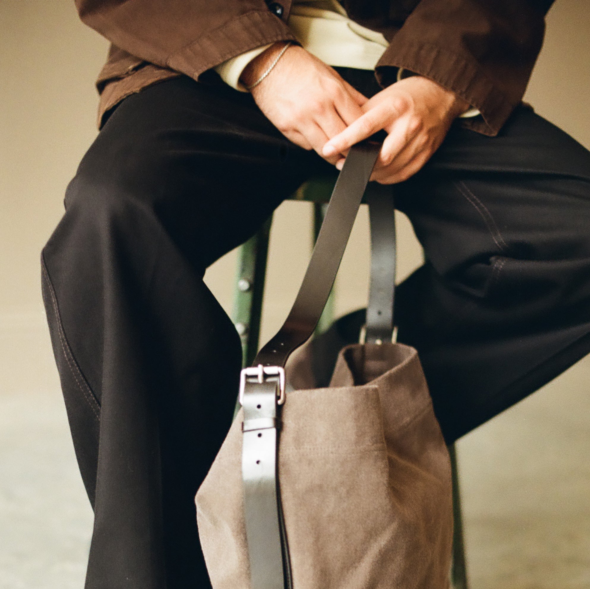A person seated and holding a large taupe canvas tote bag with dark brown leather straps and silver-tone hardware.