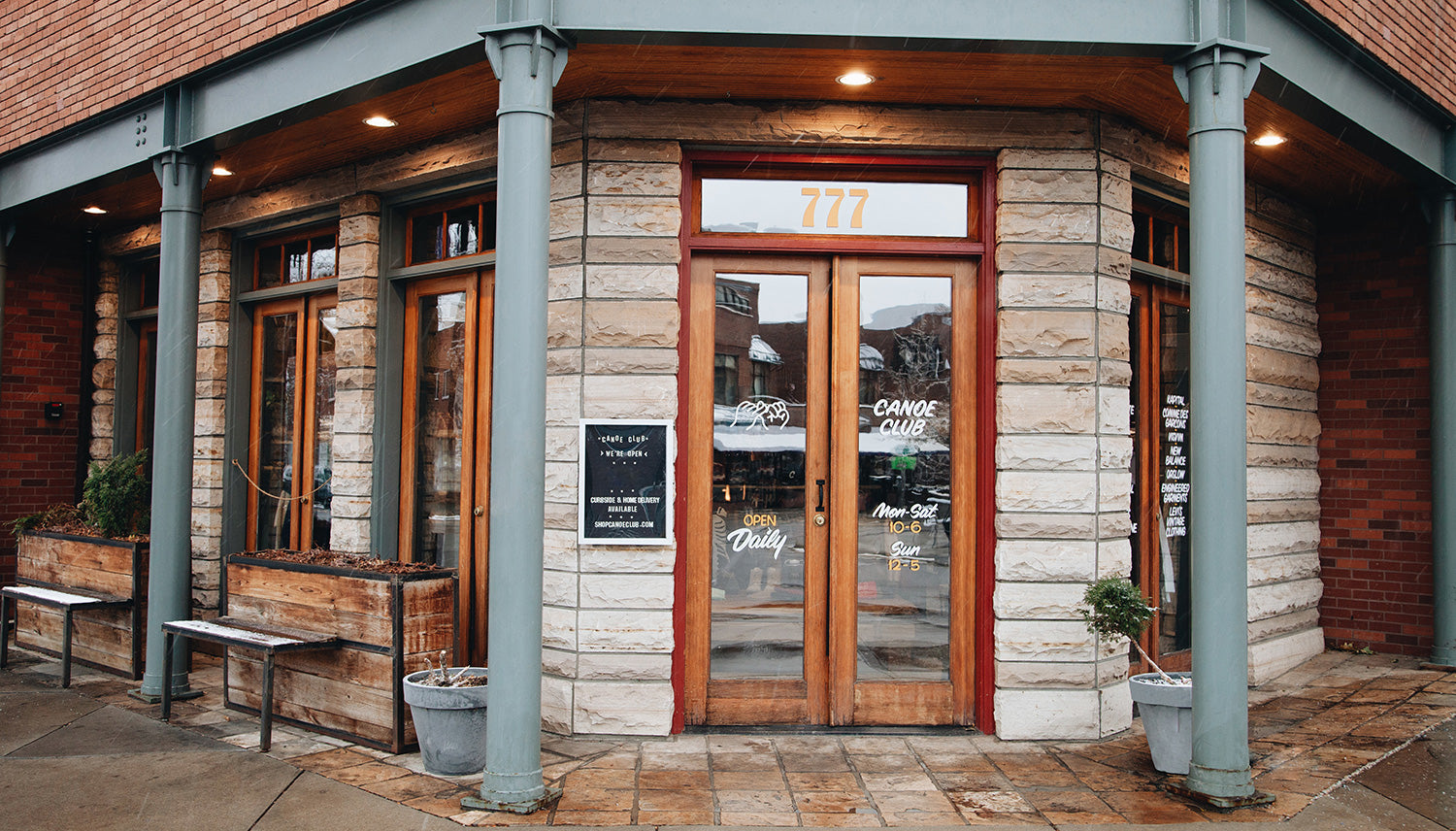 Front of Canoe Club's stone building with glass doors and wooden benches.