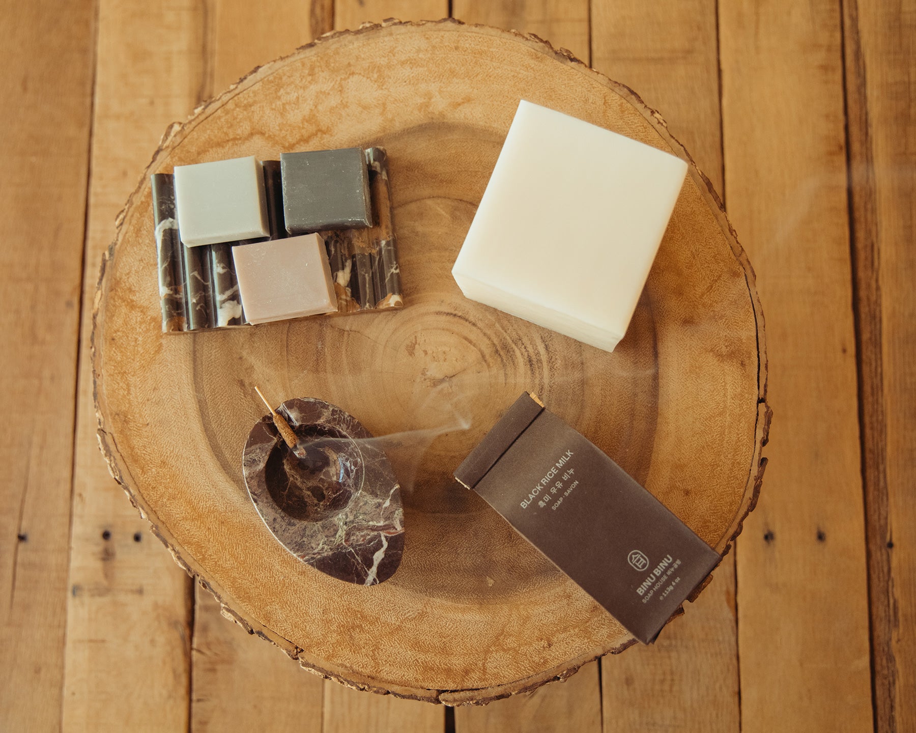 A styled flat-lay bars of soap and incense on a rustic wooden tray, set against a wooden plank background. The soap bar is off-white, accompanied by a small marble ashtray with an incense stick, and several smaller soap bars.