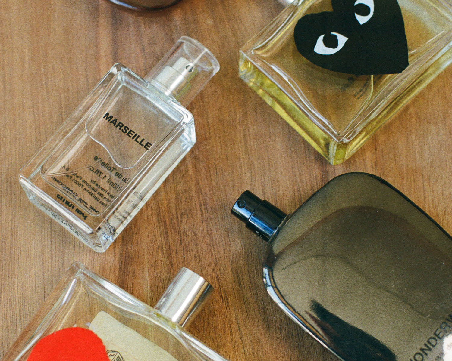 Several perfume bottles arranged on a wooden surface, including a clear rectangular bottle labeled MARSEILLE, a square bottle with a black heart-shaped label, a dark reflective bottle, and partial other bottles at the edges.