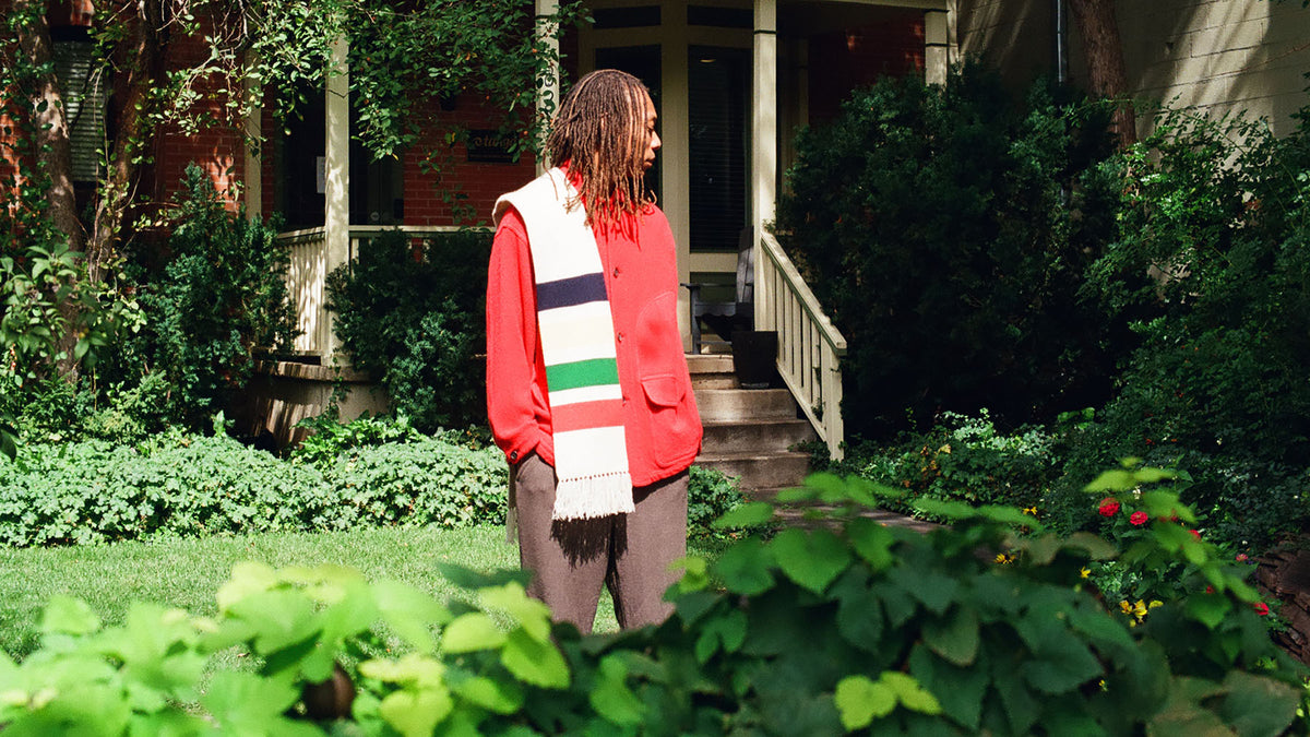 Man wearing a red button up jacket and a striped scarf standing on a lawn surrounded by greenery.