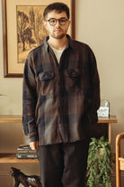 A man wearing a dark brown and black plaid flannel button-up shirt over a white t-shirt, standing in a warmly lit indoor room with framed art and wooden furniture in the background.