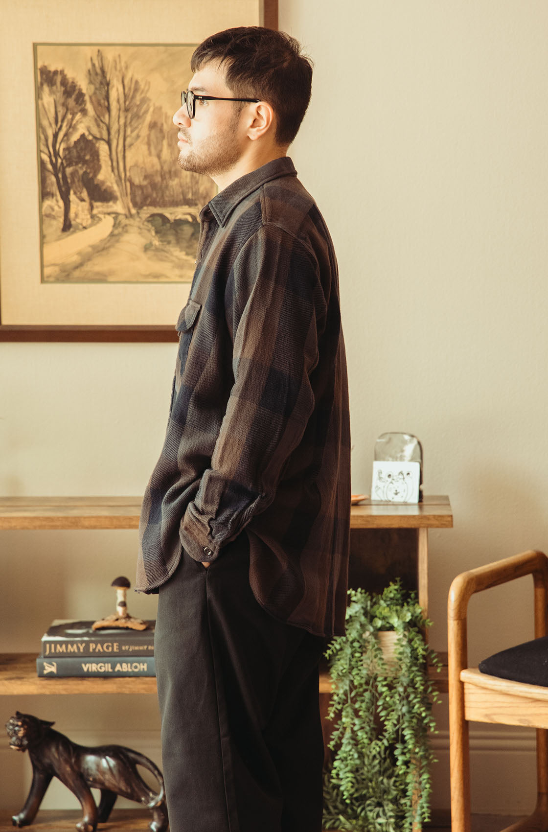 A man wearing a dark brown and black plaid flannel shirt and dark trousers, standing in profile in a warm, rustic room with a framed landscape painting on the wall.