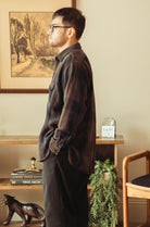 A man wearing a dark brown and black plaid flannel shirt and dark trousers, standing in profile in a warm, rustic room with a framed landscape painting on the wall.