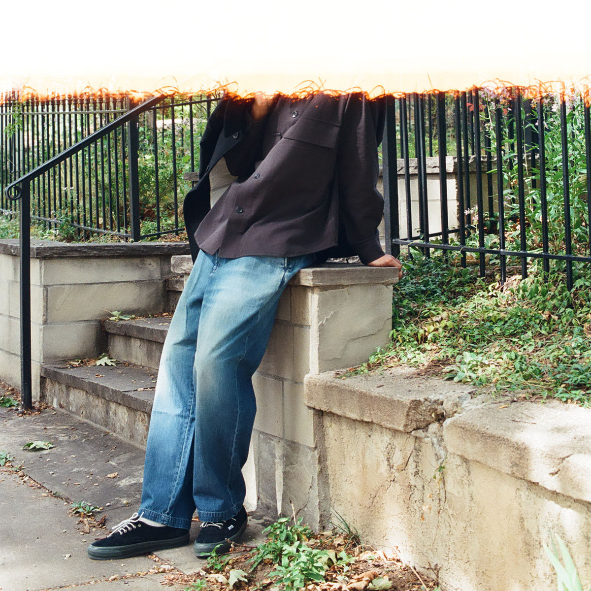 film photograph of a man, shoulders down wearing a black button up shirt, blue jeans, and black sneakers.