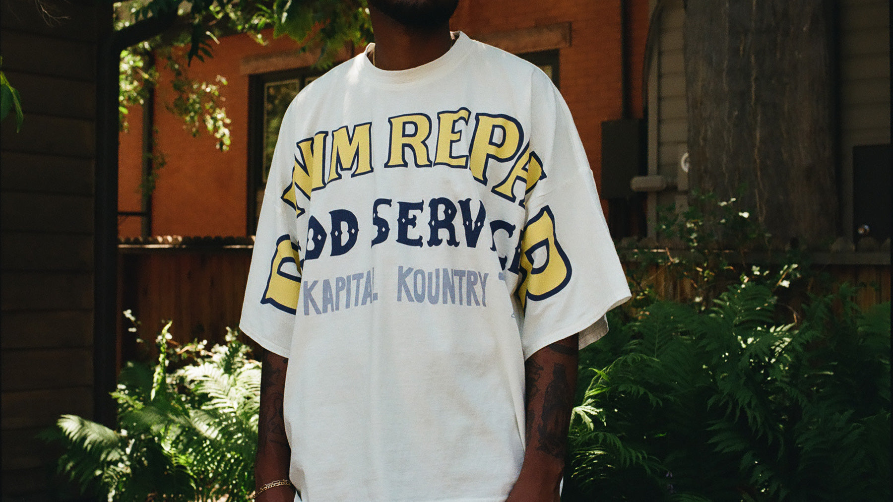 A person wearing a white graphic t-shirt with yellow and navy blue collegiate-style lettering that reads 'Denim Repair' and 'Kapital Kountry', photographed outdoors in front of green foliage and a brick building.