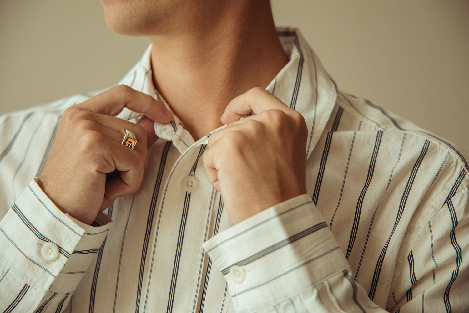 Close-up of a man adjusting the collar of a white button-down shirt with thin vertical navy stripes. He is wearing a gold ring on one finger, and the background is a soft, warm beige.