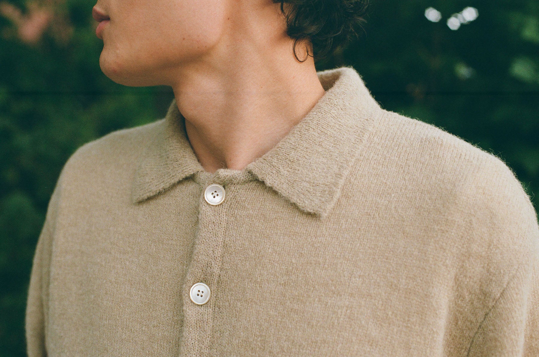 Close-up of a person wearing a soft beige knit polo shirt with a collar and white buttons, photographed outdoors against a blurred green background.