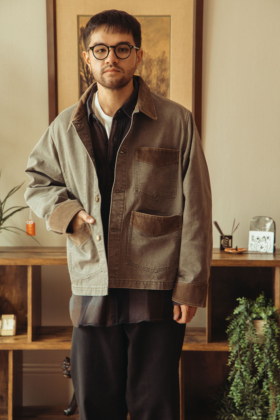 A man wearing a washed black jacket over a dark shirt and black pants, standing in a warm, indoor room with wooden shelves and framed art in the background.