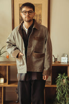 A man wearing a washed black jacket over a dark shirt and black pants, standing in a warm, indoor room with wooden shelves and framed art in the background.