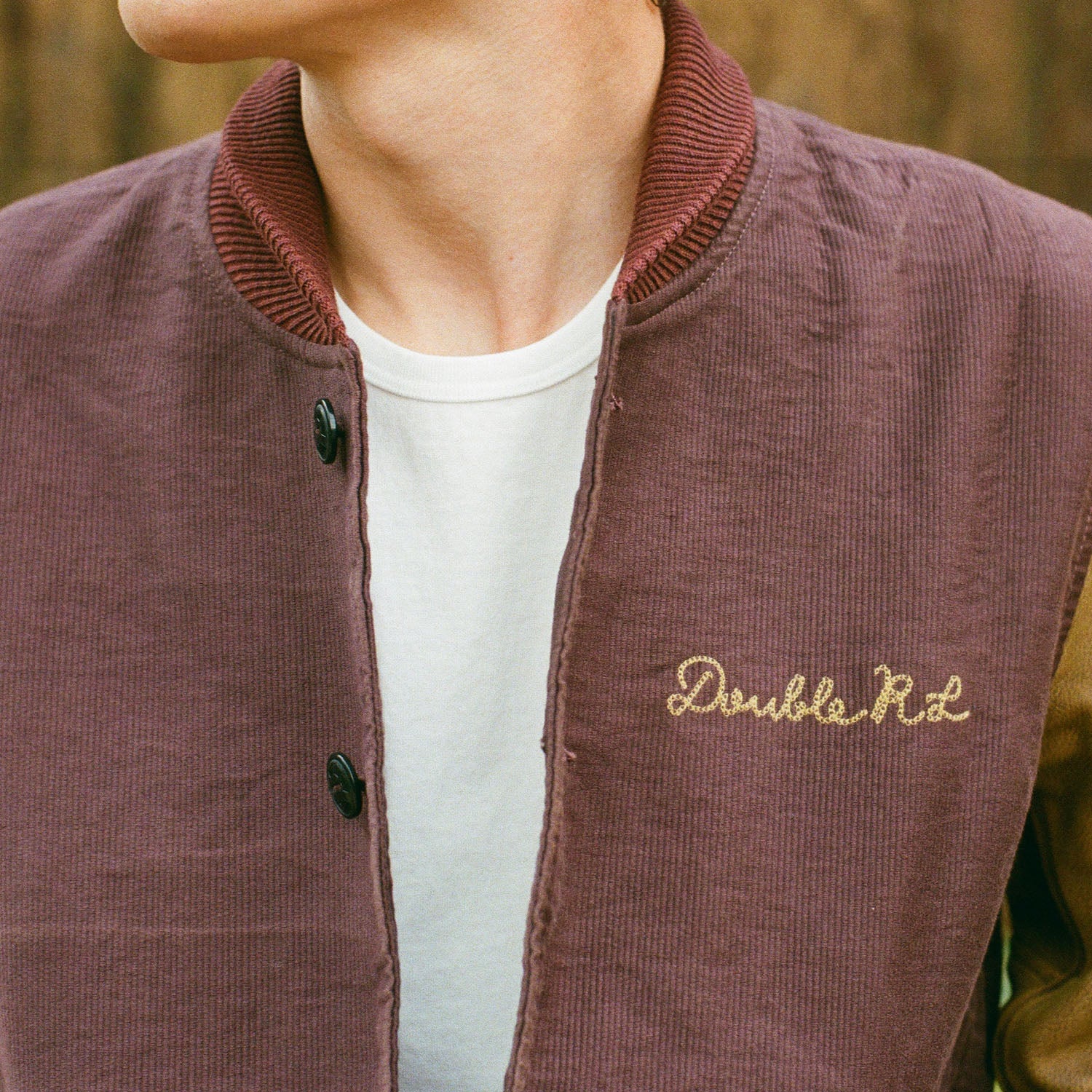 A close-up of a person wearing a corduroy varsity-style jacket in muted mauve-brown, layered over a white T-shirt. The jacket has a ribbed collar, dark buttons, and 'Double Rd' embroidered in gold on the chest.