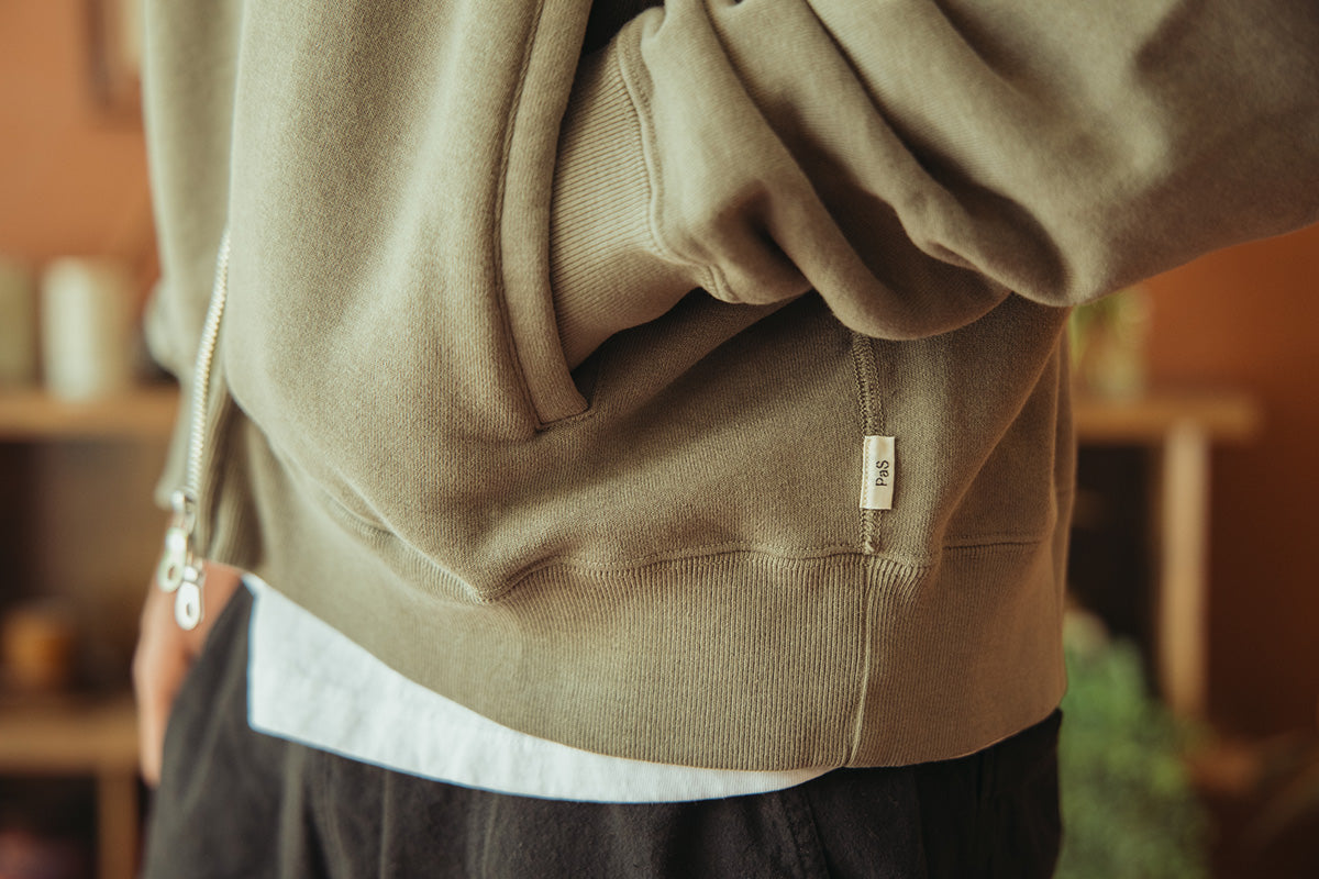 Close-up of a person wearing a taupe-beige pullover hoodie with a small white brand tag on the lower front, paired with a white undershirt and dark pants in a warm indoor setting.
