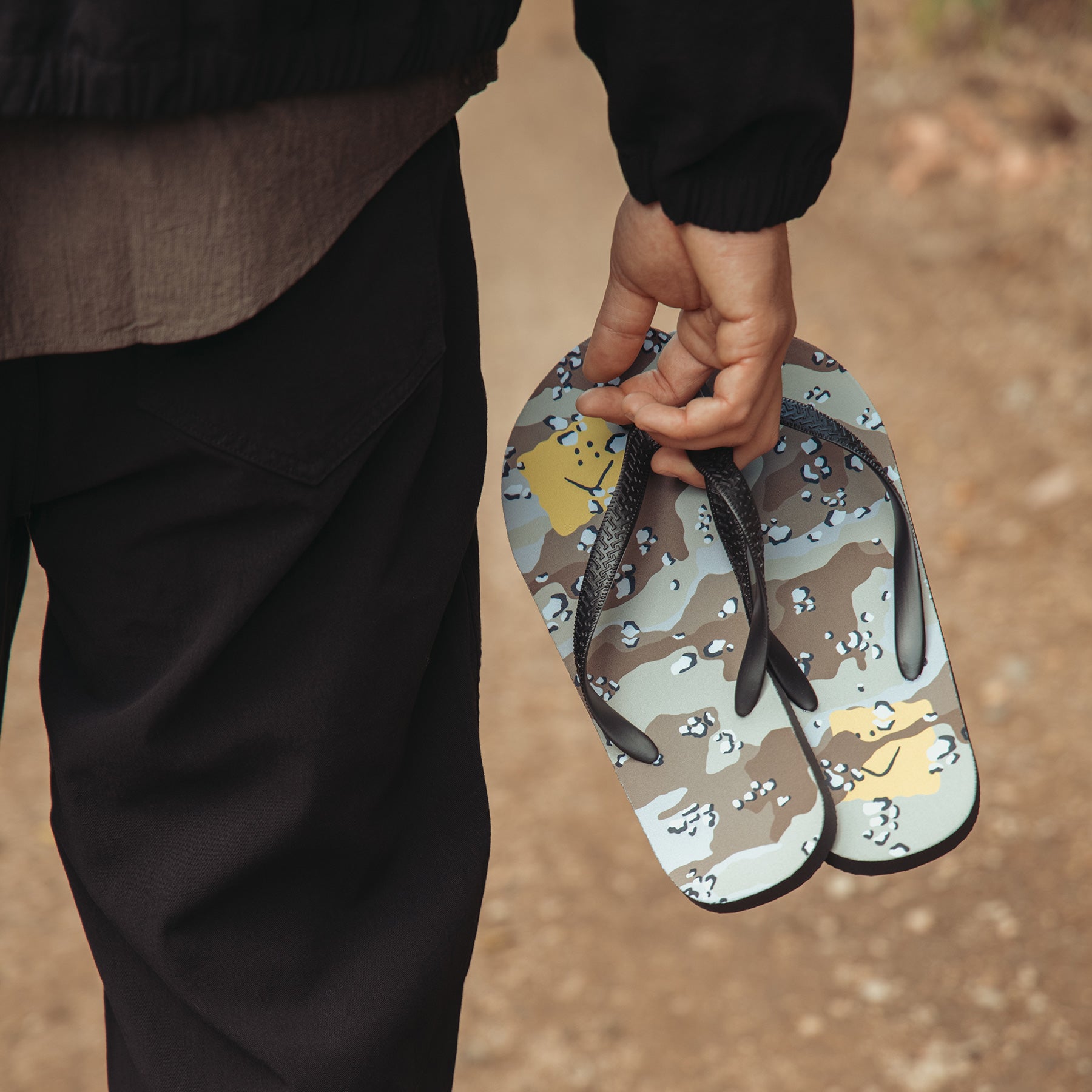 A person outdoors holding a pair of camouflage-patterned flip-flops with dark straps. The background is a blurred dirt path.