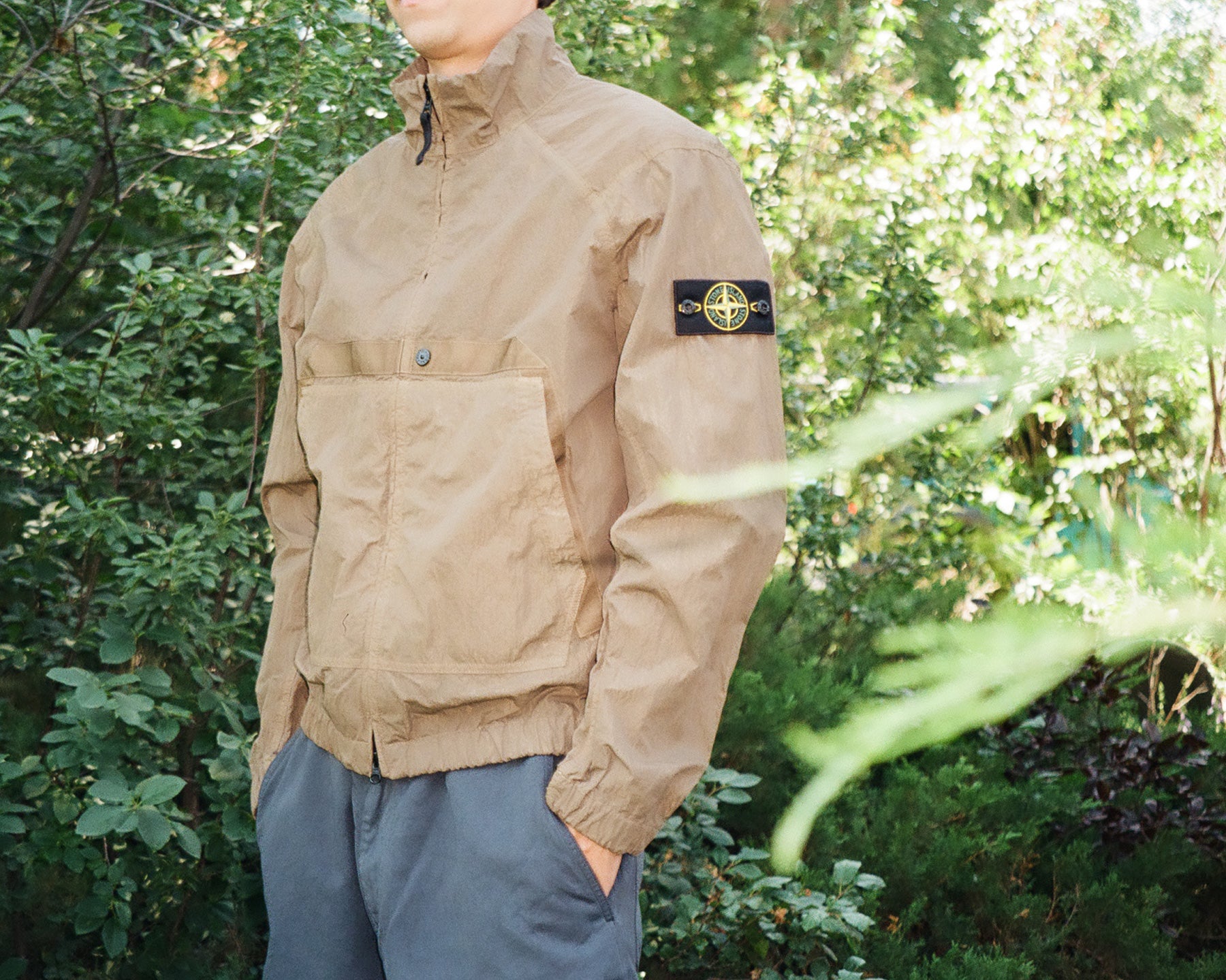 A person wearing a tan Stone Island jacket with the brand's compass patch visible on the sleeve, standing outdoors in front of green foliage.