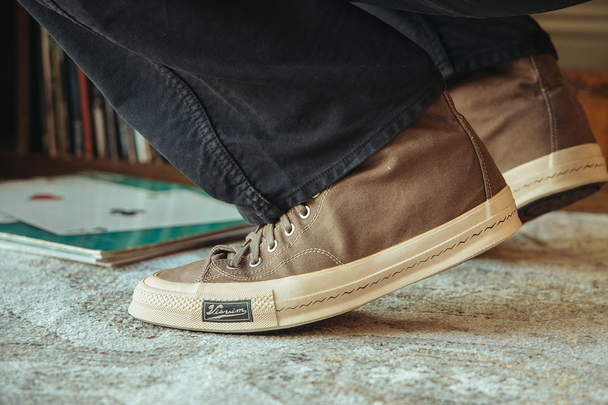 A close-up lifestyle shot of a person wearing brown canvas high-top sneakers with cream soles, standing on a textured gray carpet with a bookshelf in the background.