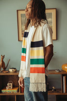 Man wearing cream scarf with multicolored stripes standing in front of a blue wall.