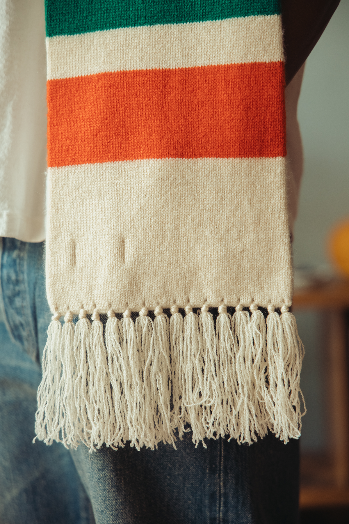Detail shot of a cream scarf with red and green stripes and a fringed bottom.