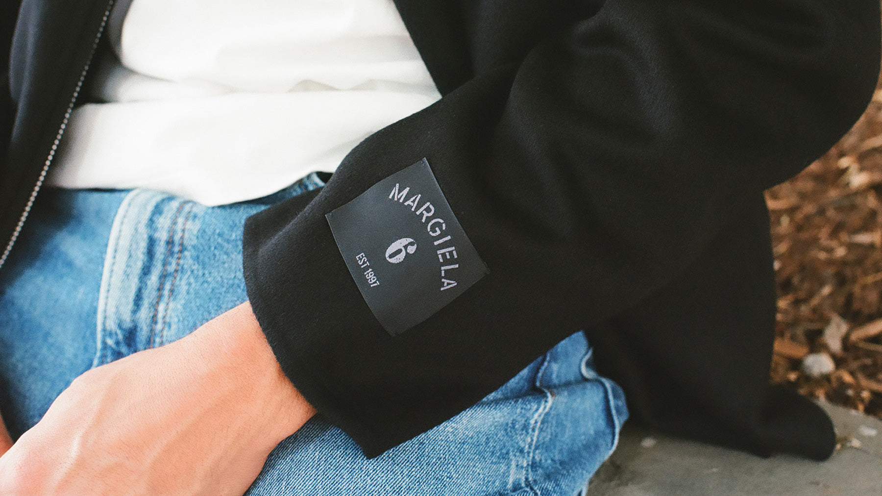 Close-up of a person wearing a black jacket and blue jeans, showing a black fabric patch on the sleeve with the brand name 'MARGIELA' and a small 'EST. 1997' label.