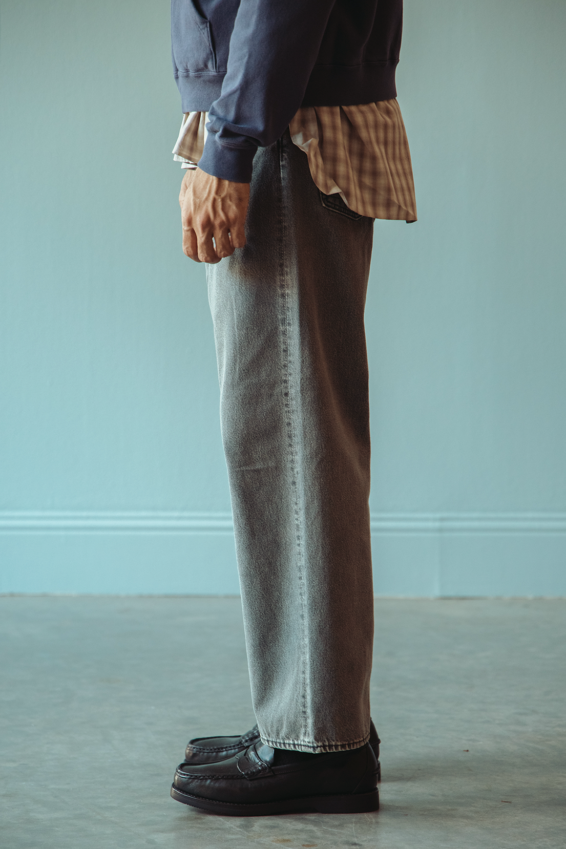 A person in profile shown from the waist down wearing light gray wide-leg denim jeans, a navy blue sweatshirt layered over a tan plaid shirt, and black leather loafers, standing against a pale blue wall.