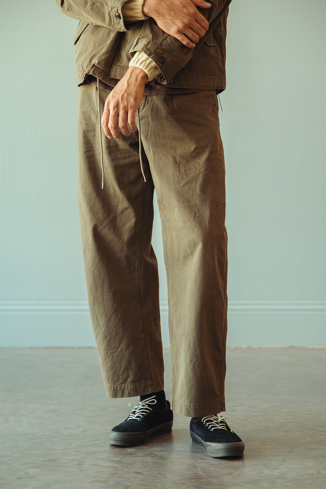 A person wearing loose, wide-leg brown pants with a drawstring waist, paired with a matching brown jacket and black low-top sneakers, standing against a pale blue-gray wall and concrete floor.