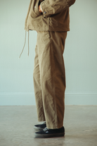 A person wearing loose brown utility pants and a matching brown jacket, standing indoors against a pale wall and concrete floor with black low-top sneakers.